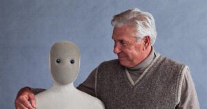 1X NEO humanoid robot in a beige knit suit beside a smiling older man in a sweater vest who has his arm around the robot
