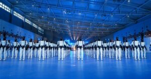 Rows of AgiBot Expedition A3 humanoid robots, white bodies with black heads, standing in formation on a blue-lit floor inside a large hall with a steel-truss roof