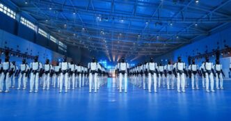 Rows of AgiBot Expedition A3 humanoid robots, white bodies with black heads, standing in formation on a blue-lit floor inside a large hall with a steel-truss roof