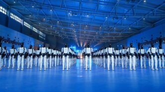 Rows of AgiBot Expedition A3 humanoid robots, white bodies with black heads, standing in formation on a blue-lit floor inside a large hall with a steel-truss roof