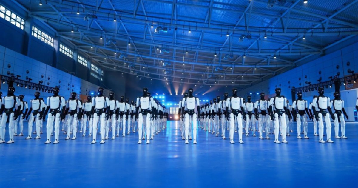 Rows of AgiBot Expedition A3 humanoid robots, white bodies with black heads, standing in formation on a blue-lit floor inside a large hall with a steel-truss roof