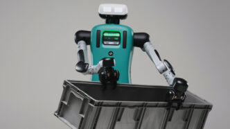Agility Robotics Digit humanoid robot with a teal torso carrying a grey plastic tote against a plain grey background