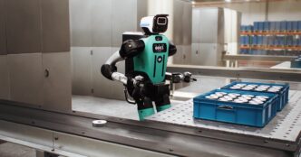 Agility Robotics' teal and black Digit 5 humanoid with a white head standing at a factory workbench beside two blue totes of metal parts