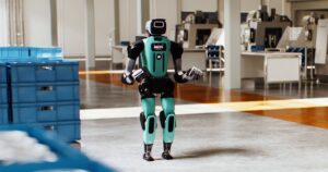 Agility's teal and black Digit 5 humanoid standing on a factory floor beside stacked blue totes, in a still from Agility's Introducing Digit 5 video