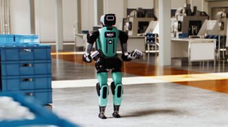 Agility's teal and black Digit 5 humanoid standing on a factory floor beside stacked blue totes, in a still from Agility's Introducing Digit 5 video