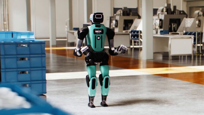 Agility's teal and black Digit 5 humanoid standing on a factory floor beside stacked blue totes, in a still from Agility's Introducing Digit 5 video