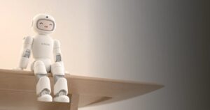 AiMOGA's Mobo, a small white humanoid home robot with a rounded helmet-like head and a smiling face on its display, sitting on the edge of a wooden table against a warm beige wall