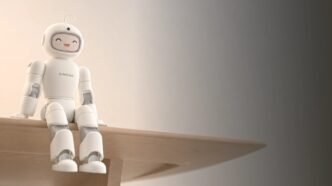 AiMOGA's Mobo, a small white humanoid home robot with a rounded helmet-like head and a smiling face on its display, sitting on the edge of a wooden table against a warm beige wall