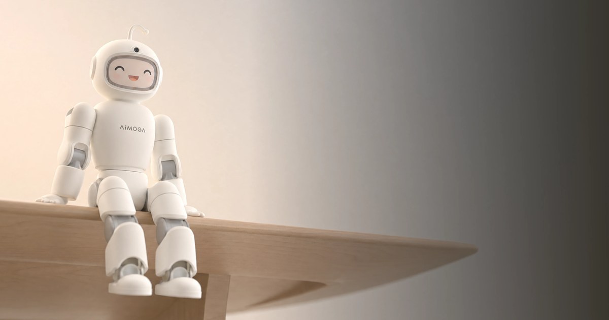 AiMOGA's Mobo, a small white humanoid home robot with a rounded helmet-like head and a smiling face on its display, sitting on the edge of a wooden table against a warm beige wall