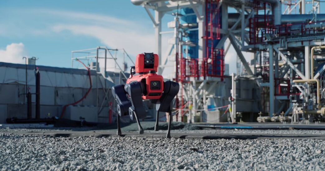 A red and dark blue four-legged ANYmal X inspection robot walking on gravel in front of steel pipework and a processing tower at an industrial plant under a blue sky