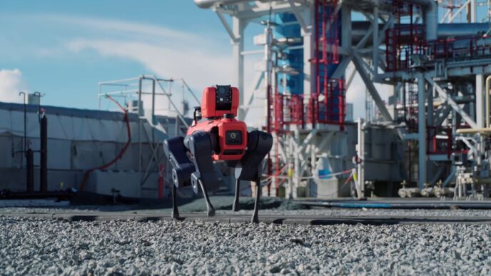 A red and dark blue four-legged ANYmal X inspection robot walking on gravel in front of steel pipework and a processing tower at an industrial plant under a blue sky