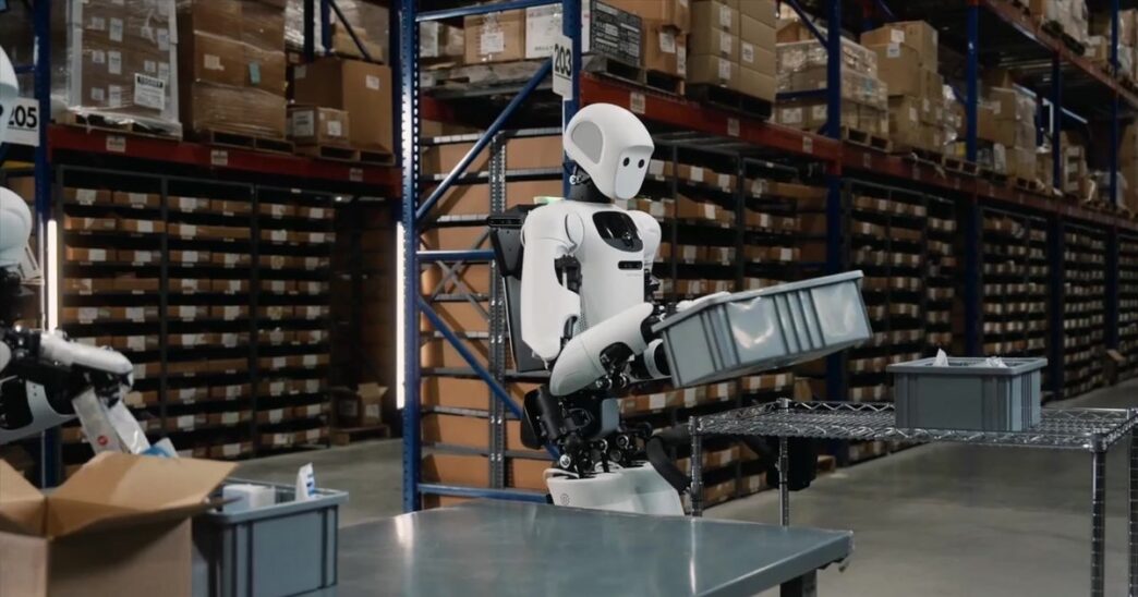Apptronik Apollo humanoid robot lifting a grey tote in a warehouse aisle lined with shelving