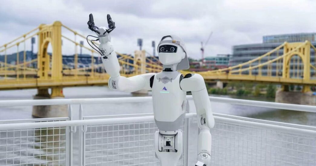 White Astribot T1 robot with a black visor-style face and a cap, one gripper raised, at a riverside railing in front of a yellow suspension bridge