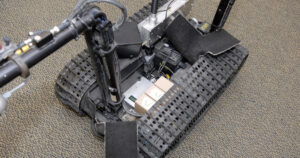 Top-down view of a tracked TALON bomb-disposal robot with its equipment bay open, showing two tan modules, wiring and electronics between black rubber tracks on a grey carpet