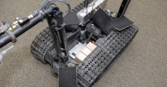Top-down view of a tracked TALON bomb-disposal robot with its equipment bay open, showing two tan modules, wiring and electronics between black rubber tracks on a grey carpet