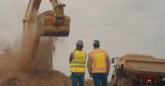Two construction workers in hard hats and yellow high-visibility vests, one marked Bedrock, watching an excavator bucket drop dirt beside a yellow haul truck under a grey sky