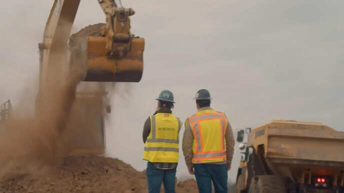 Two construction workers in hard hats and yellow high-visibility vests, one marked Bedrock, watching an excavator bucket drop dirt beside a yellow haul truck under a grey sky