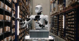 Apptronik Apollo humanoid robot in a warehouse aisle reaching toward shelving while holding a barcode scanner, head visible