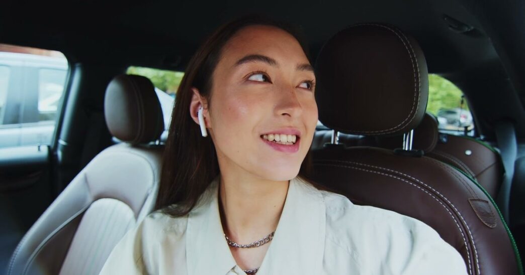 A smiling young woman wearing a wireless earbud sits in the back seat of a car with brown leather seats and looks over her shoulder as she talks