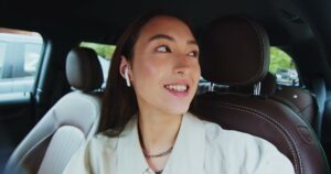 A smiling young woman wearing a wireless earbud sits in the back seat of a car with brown leather seats and looks over her shoulder as she talks