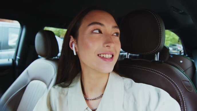 A smiling young woman wearing a wireless earbud sits in the back seat of a car with brown leather seats and looks over her shoulder as she talks
