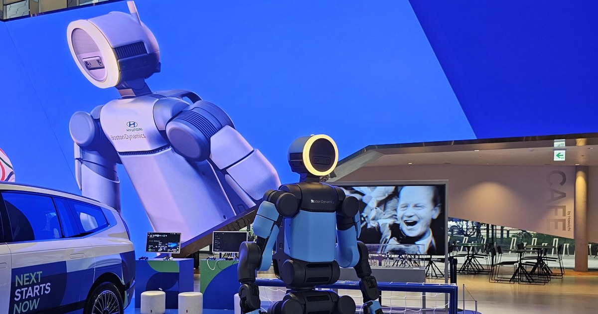 Boston Dynamics' blue-and-black electric Atlas humanoid with a ring-lit head, seen from behind in front of a giant LED-wall image of Atlas at Hyundai Motorstudio Goyang