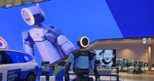 Boston Dynamics' electric Atlas humanoid with a ring-lit head on a display platform at Hyundai Motorstudio Goyang, beside a Hyundai car and in front of a giant LED-wall image of Atlas