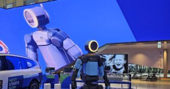 Boston Dynamics' electric Atlas humanoid with a ring-lit head on a display platform at Hyundai Motorstudio Goyang, beside a Hyundai car and in front of a giant LED-wall image of Atlas