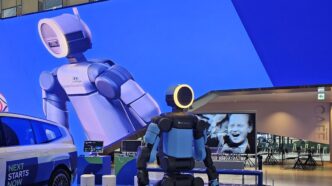 Boston Dynamics' electric Atlas humanoid with a ring-lit head on a display platform at Hyundai Motorstudio Goyang, beside a Hyundai car and in front of a giant LED-wall image of Atlas