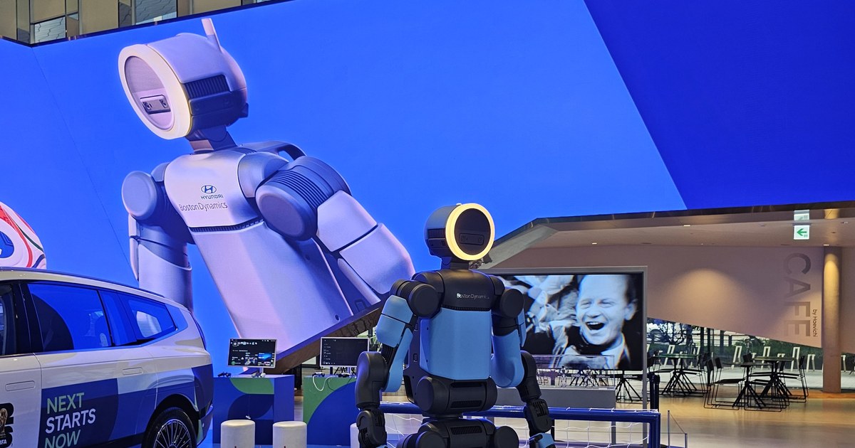 Boston Dynamics' electric Atlas humanoid with a ring-lit head on a display platform at Hyundai Motorstudio Goyang, beside a Hyundai car and in front of a giant LED-wall image of Atlas
