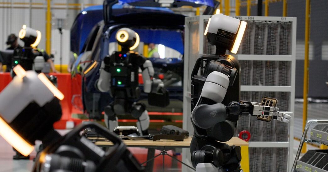 Several Boston Dynamics Atlas humanoid robots with glowing ring-lit heads handling parts at a work table beside a blue car body and parts racks, with a worker wearing a VR headset in the background
