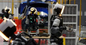 Several Boston Dynamics Atlas humanoid robots with glowing ring-lit heads handling parts at a work table beside a blue car body and parts racks, with a worker wearing a VR headset in the background