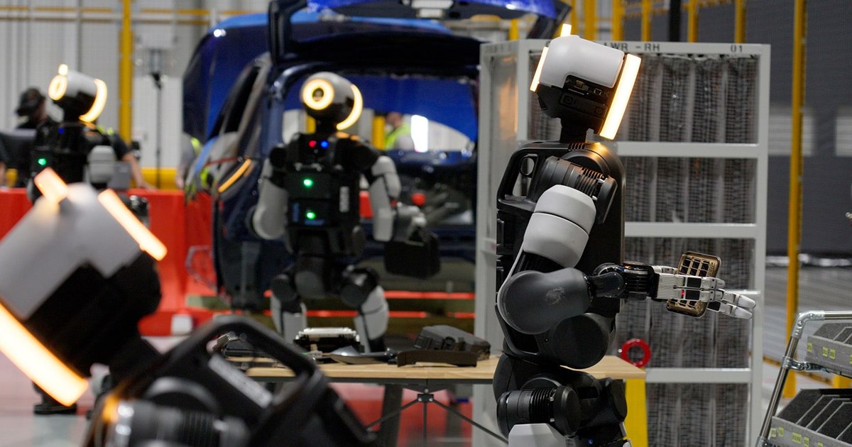 Several Boston Dynamics Atlas humanoid robots with glowing ring-lit heads handling parts at a work table beside a blue car body and parts racks, with a worker wearing a VR headset in the background