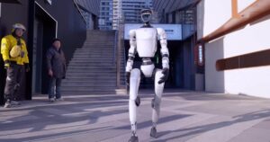 Unitree G1 humanoid robot walking across a city plaza while two pedestrians watch from a doorway