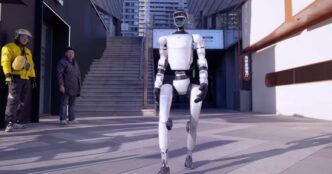 Unitree G1 humanoid robot walking across a city plaza while two pedestrians watch from a doorway