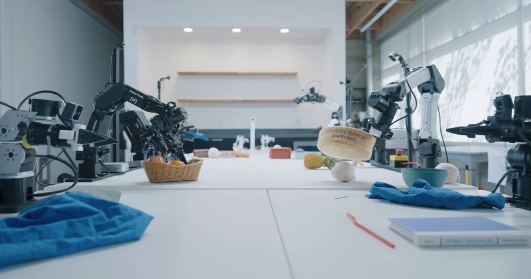 Robot arms from several makers on long white lab tables at Dream Labs, one gripper lifting a container, in a still from the Dream-1 launch video