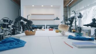 Robot arms from several makers on long white lab tables at Dream Labs, one gripper lifting a container, in a still from the Dream-1 launch video