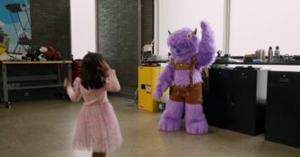 Hugo, a purple furry character robot with small horns and embroidered brown lederhosen, raising one arm to wave at a little girl in a pink tutu in Dynamic Creatures' workshop