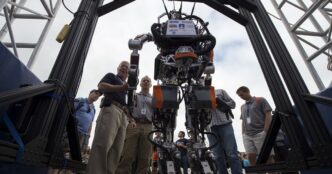 Virginia Tech's ESCHER humanoid robot standing upright inside a heavy black support frame outdoors while engineers gather around it at the DARPA Robotics Challenge