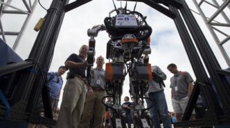 Virginia Tech's ESCHER humanoid robot standing upright inside a heavy black support frame outdoors while engineers gather around it at the DARPA Robotics Challenge