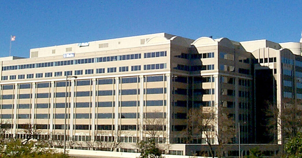 Exterior of the Federal Communications Commission building, a tan multi-storey office building with rooftop flags and bare trees under a clear blue sky