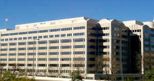 Exterior of the Federal Communications Commission building, a tan multi-storey office building with rooftop flags and bare trees under a clear blue sky