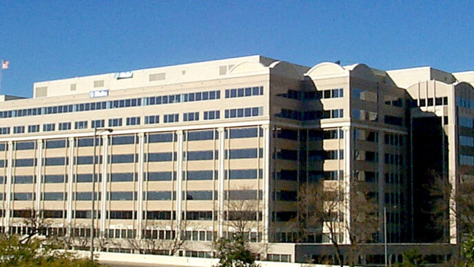 Exterior of the Federal Communications Commission building, a tan multi-storey office building with rooftop flags and bare trees under a clear blue sky