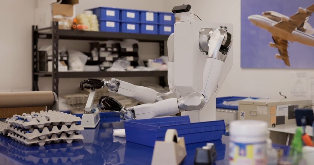 White two-armed Feather robot on a wheeled torso reaching across a blue workbench in a parts workshop