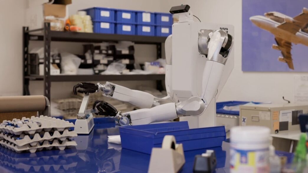 White two-armed Feather robot on a wheeled torso reaching across a blue workbench in a parts workshop
