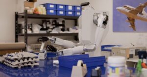 White two-armed Feather robot on a wheeled torso reaching across a blue workbench in a parts workshop