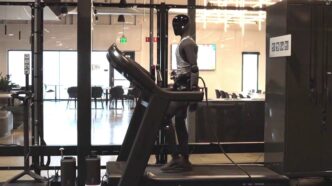 Figure 03 humanoid robot in a gray tank top walking on a treadmill in Figure's office, with a cable running from the robot