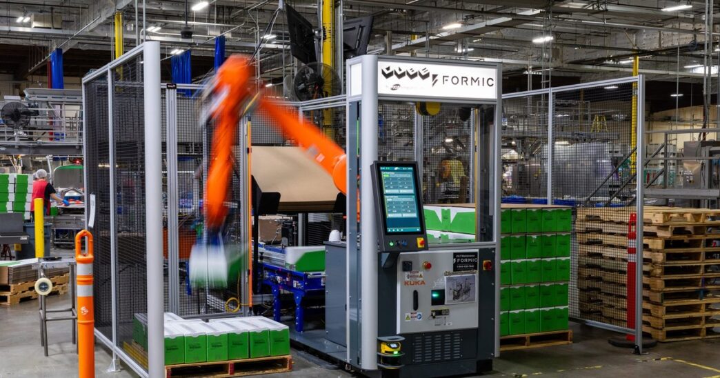 An orange KUKA robot arm, blurred in motion, stacking green cases inside a fenced Formic palletizing cell with a touchscreen control cabinet, on a warehouse floor beside stacked wooden pallets