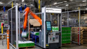 An orange KUKA robot arm, blurred in motion, stacking green cases inside a fenced Formic palletizing cell with a touchscreen control cabinet, on a warehouse floor beside stacked wooden pallets