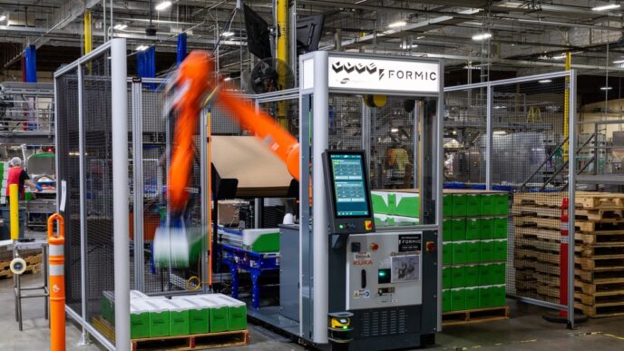 An orange KUKA robot arm, blurred in motion, stacking green cases inside a fenced Formic palletizing cell with a touchscreen control cabinet, on a warehouse floor beside stacked wooden pallets